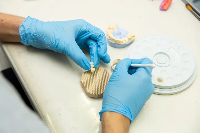 Gloved hands at a dental laboratory bench, seen from above, working on a set of artificial teeth.