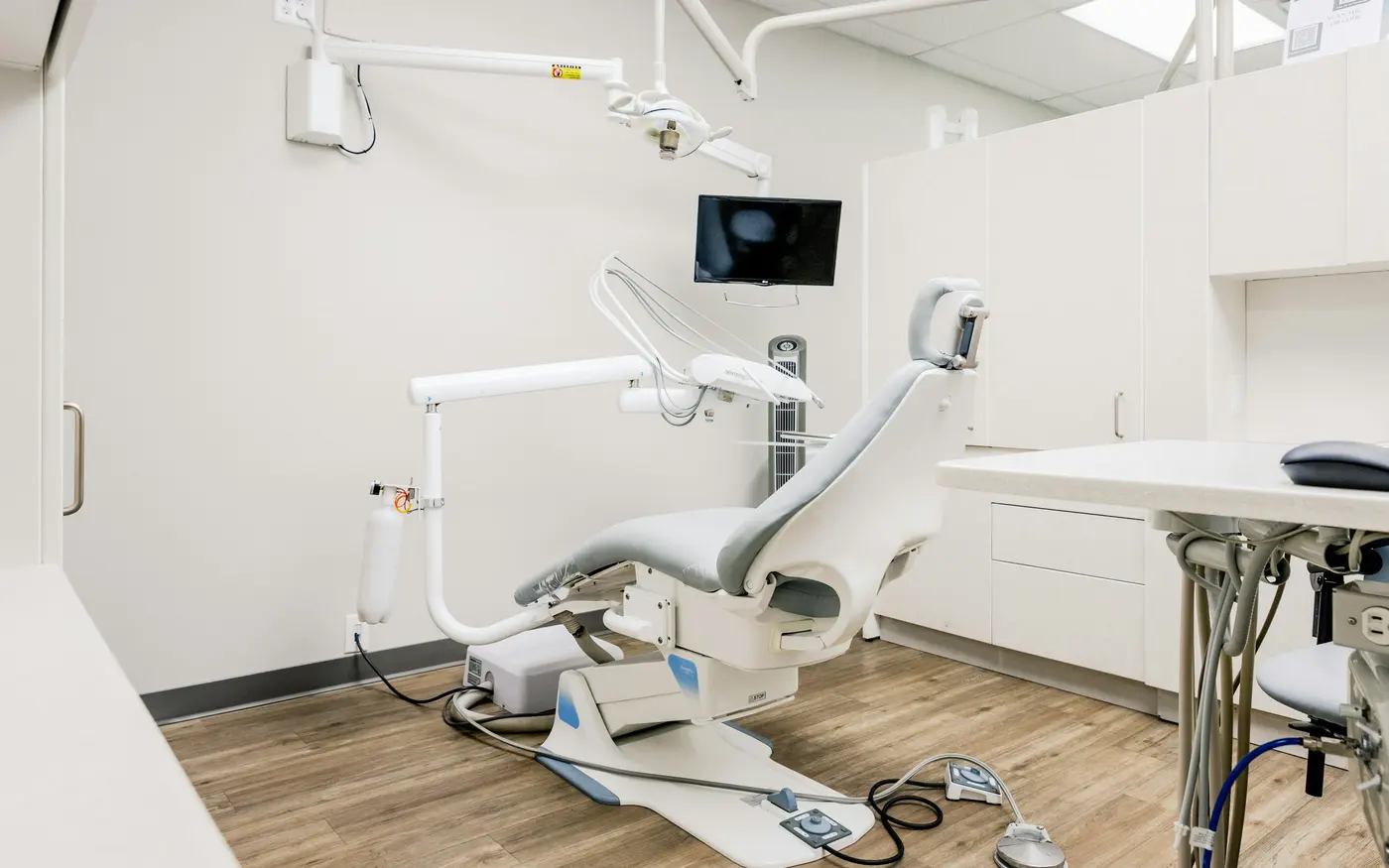 An empty dental treatment room with a pale chair, wood floor and daylight from a tall window.